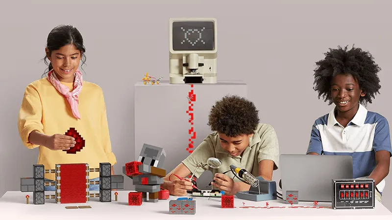 Three students building redstone contraptions at a table with Minecraft blocks
